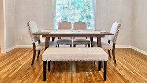 A dining room with a wooden table and six upholstered chairs, a bench, and a large window with blinds. - Tranquil Ranch Style Retreat w/ 2+ Acres of Land (Marietta)