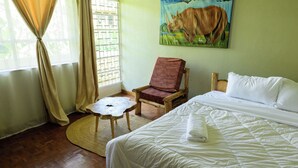 A hotel room with a bed, a chair, a small table, a painting of a cow, and a window with curtains. - 5-bedroom bed & breakfast in charming Arusha with WiFi (Arusha)