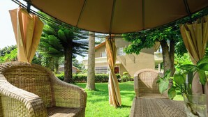 A covered outdoor seating area with wicker furniture and a glass table, surrounded by greenery and a house in the background. - 5-bedroom bed & breakfast in charming Arusha with WiFi (Arusha)