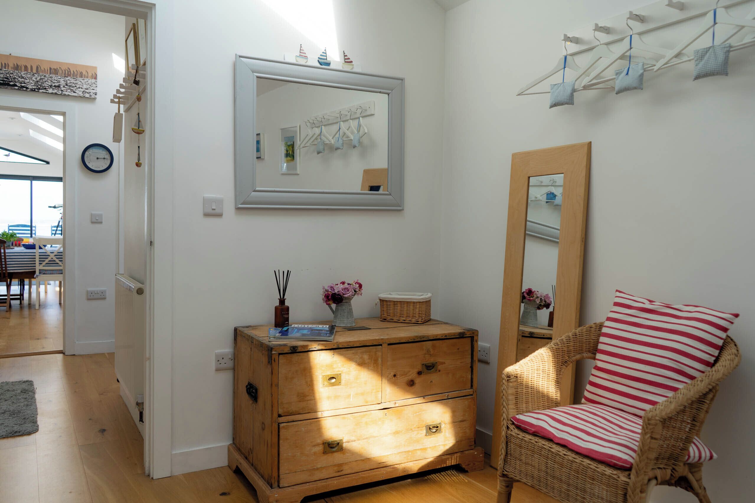 Una habitación con un aparador de madera, una silla de mimbre con cojín a rayas, un espejo y un tendedero de pared.