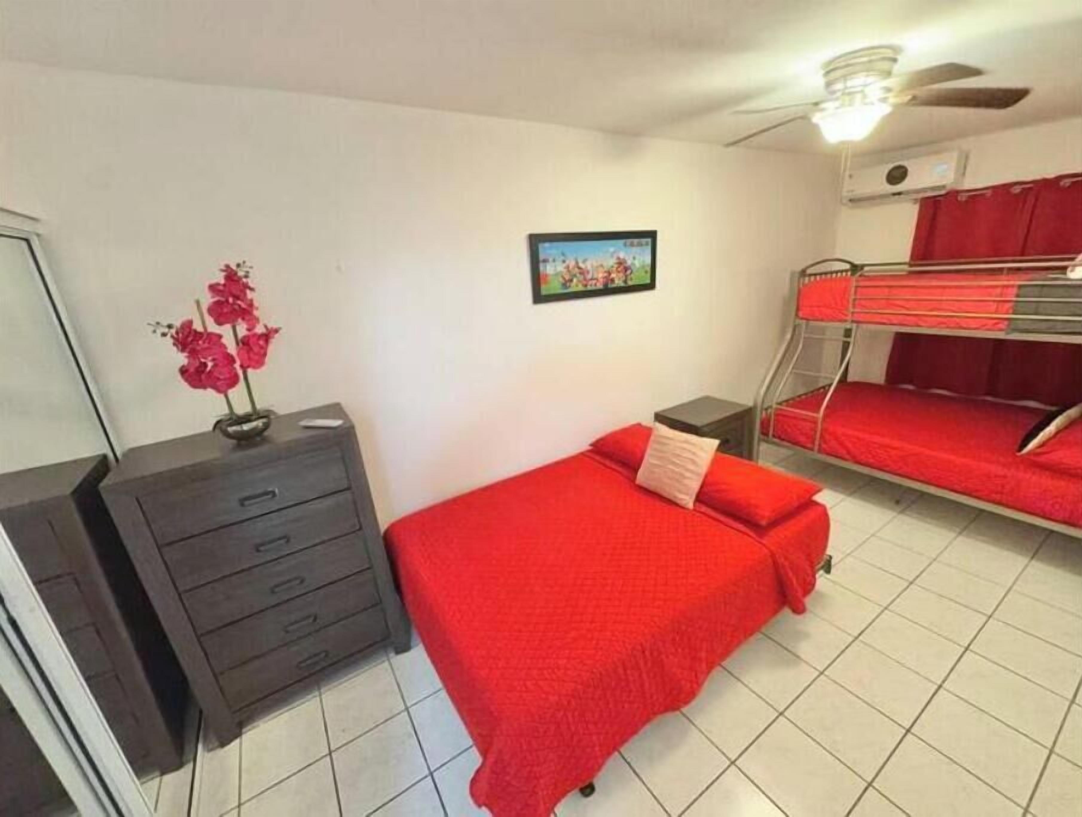 Ein Zimmer mit Etagenbett, Kommode, kleinem Tisch, einem Bild an der Wand und einem Deckenventilator.