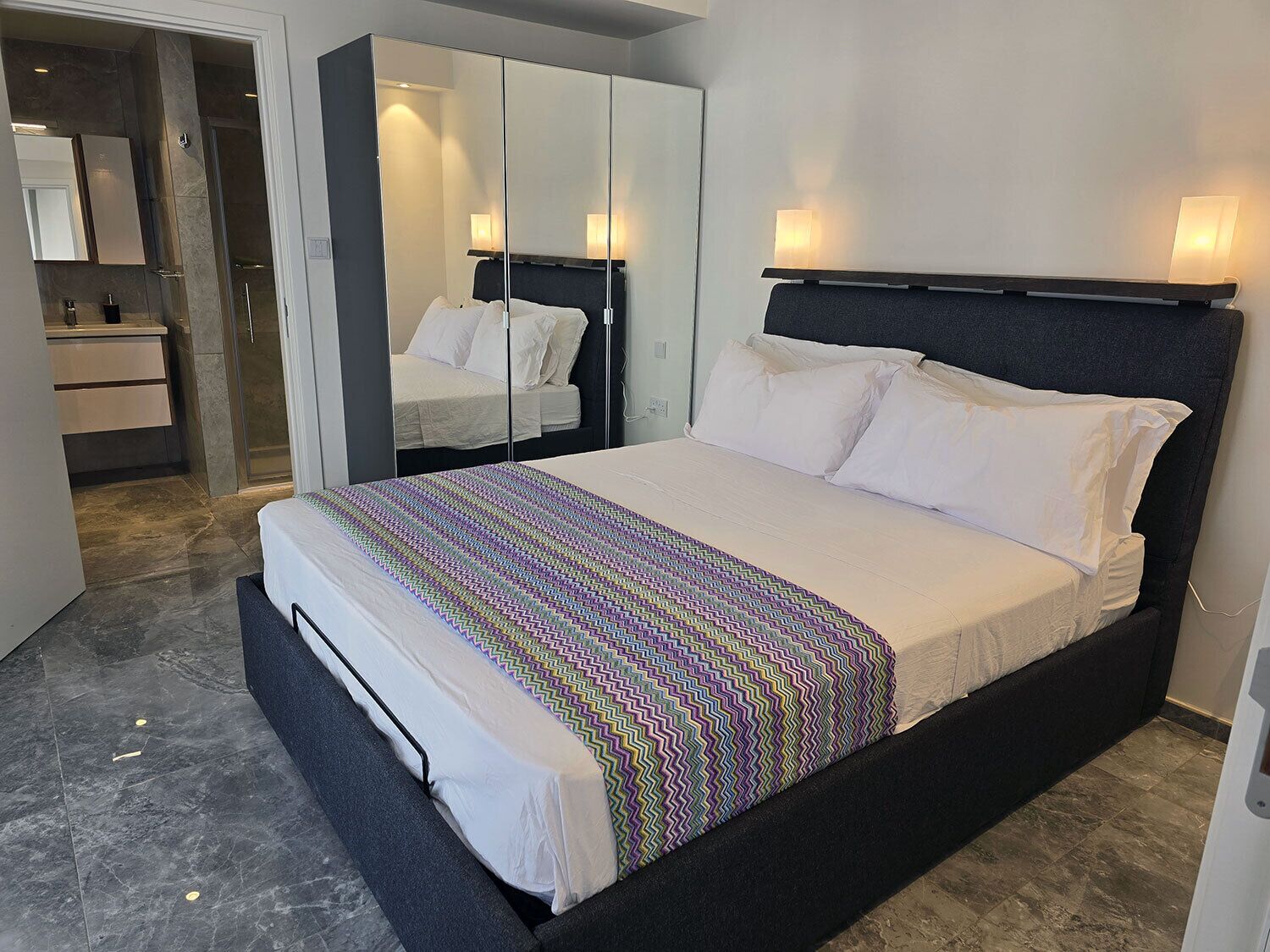 Une chambre moderne avec un grand lit, une armoire avec des miroirs, une salle de bain avec douche et un petit coin salon.