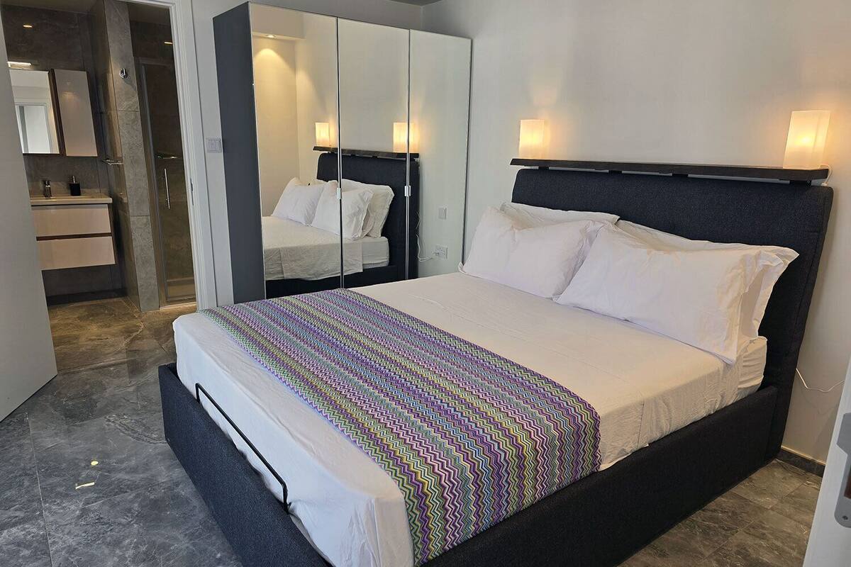 A modern bedroom with a large bed, a wardrobe with mirrors, a bathroom with a shower, and a small seating area.