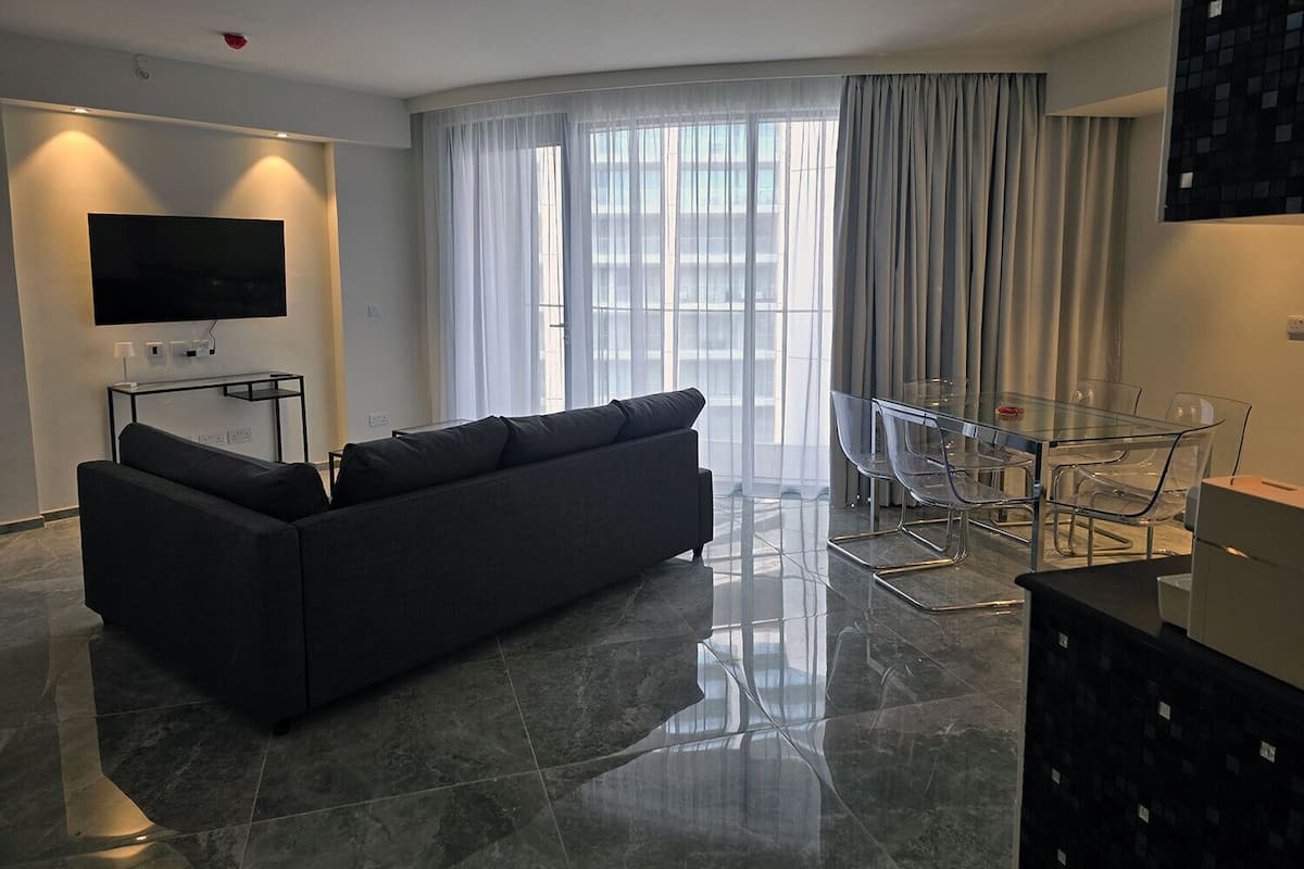 A modern living room with a dark sofa, a glass dining table, and a TV mounted on the wall.