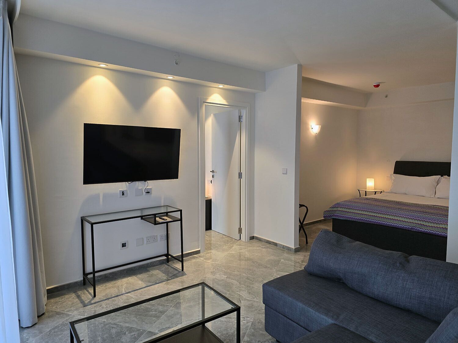 Une chambre d’hôtel moderne, dotée d’un téléviseur à écran plat fixé au mur, d’une table basse en verre, d’un canapé gris, d’un lit recouvert d’une couvre-lit à motifs et d’une porte donnant sur une autre pièce.