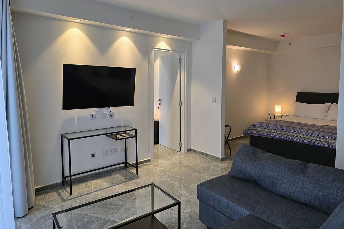 A modern hotel room with a flat-screen TV mounted on the wall, a glass coffee table, a gray sofa, a bed with a patterned bedspread, and a door leading to another room.