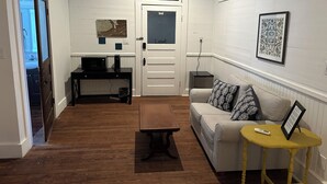 Un espace de vie compact doté d’un plancher en bois, d’un canapé avec des coussins à motifs, d’une petite table, d’un bureau avec un ordinateur et d’une porte donnant sur une autre pièce.