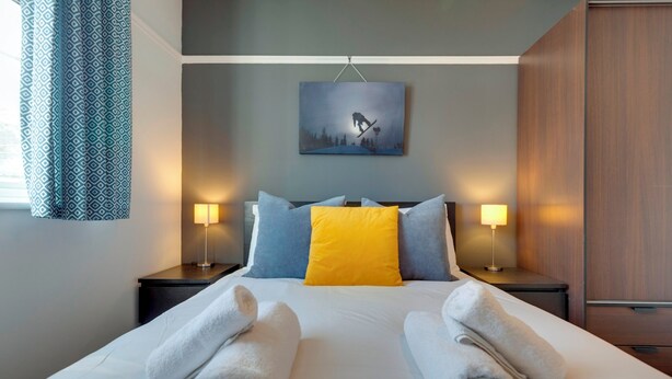 A neatly made bed with white and yellow pillows, two bedside lamps, and a painting of a snowboarder on the wall. - Stylish and Modern Flat with Free Parking | Pass the Keys (Cambridge)