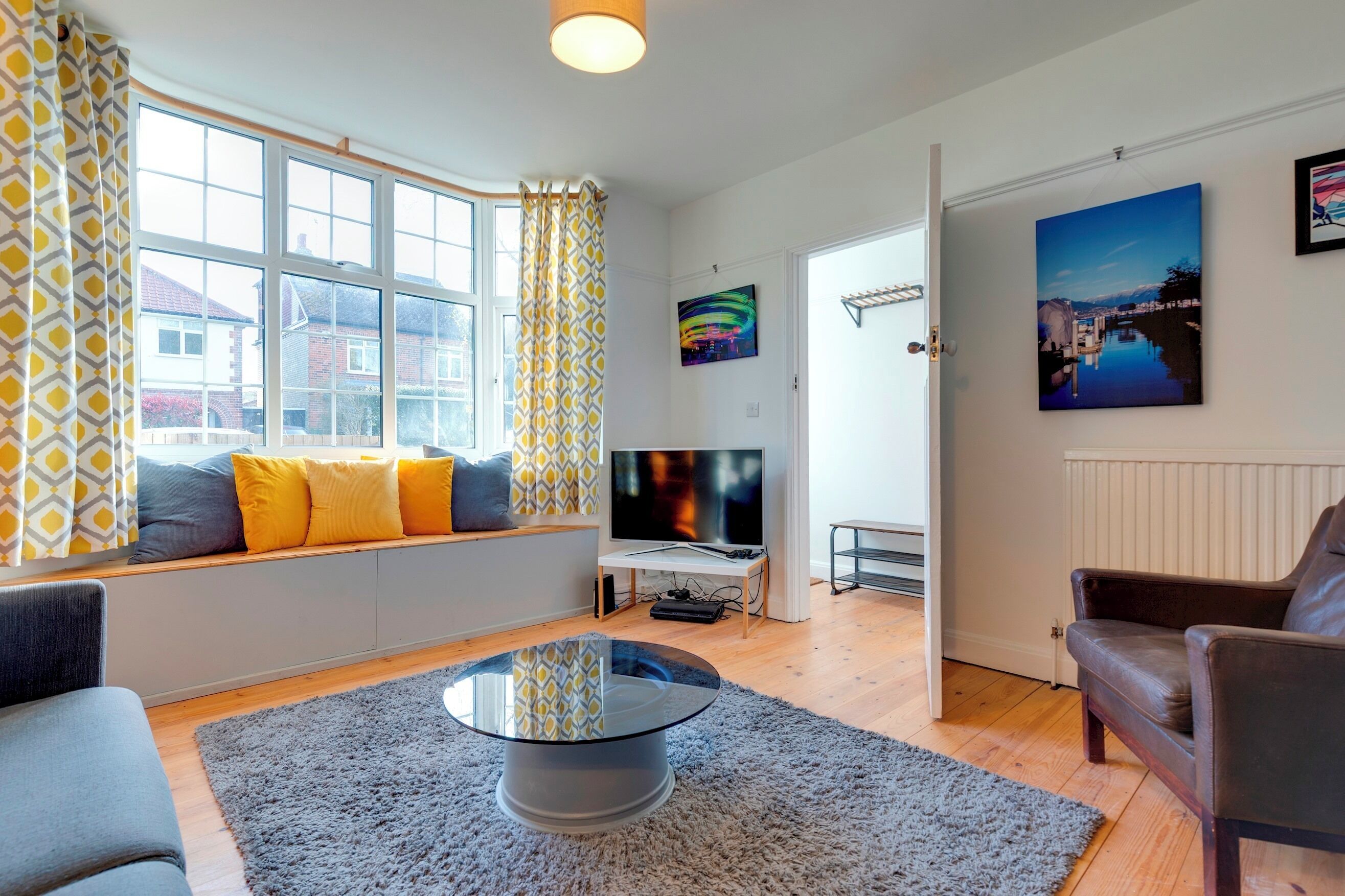 A modern living room with a sofa, a glass coffee table, and a TV. There are curtains, a painting, and a window with a view of buildings.