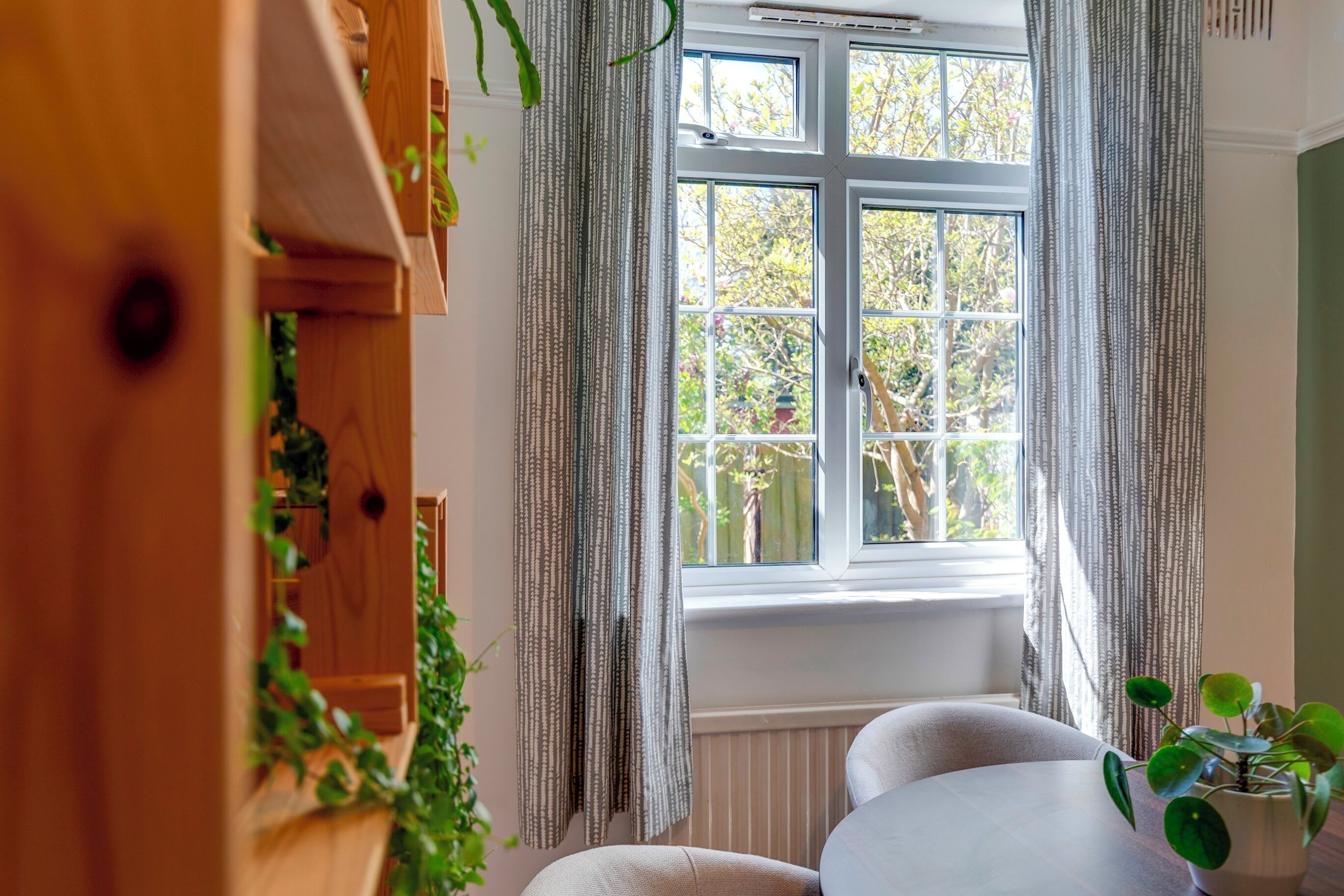 Ein Zimmer mit großem Fenster, einem Holzschrank mit Pflanzen und einem runden Tisch.