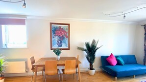 A dining area with a wooden table and chairs, a potted plant, a blue sofa with colorful cushions, and a framed artwork on the wall.