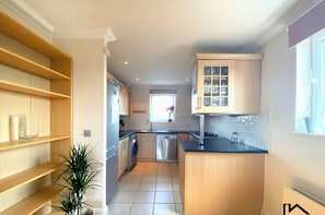 A kitchen with wooden shelves, a sink, a dishwasher, and a window with a plant.