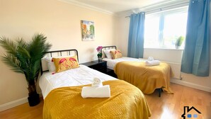 A hotel room with two beds, a potted plant, a painting on the wall, and a window with curtains.
