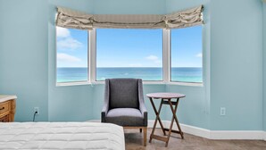 A bedroom with a large window overlooking the ocean, a gray armchair, a small round table, and a bed with a white comforter. - Tidewater 408 (Panama City Beach)