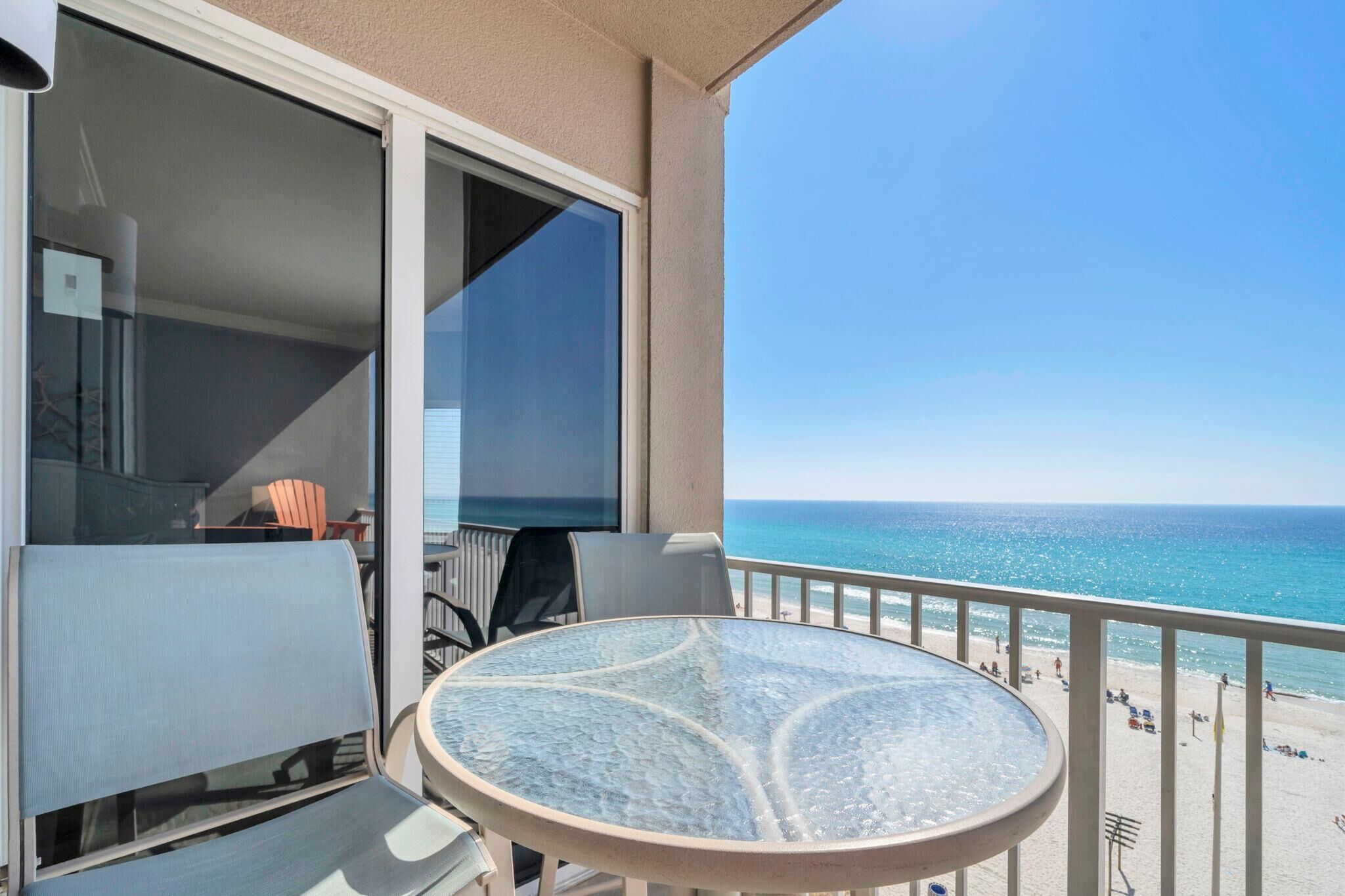 A balcony with a glass table and chairs, overlooking a beach with people and a clear sky.