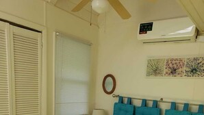 Een slaapkamer met een bed, een plafondventilator, een airconditioning, een nachtkastje, een lamp, een raam met jaloezieën, een wandspiegel en een decoratief wandobject.