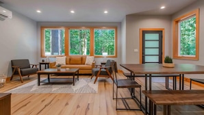 Una sala de estar moderna con sofá, mesa de centro y mesa de comedor. Ventanas amplias ofrecen luz natural y vistas a árboles.