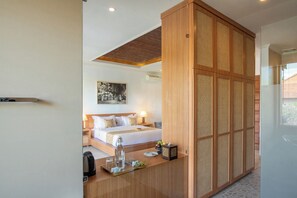 A modern hotel room with a bed, bedside lamps, a wooden desk with a tray of items, and a large wooden partition. - Budhi Ayu Suite (Ubud)