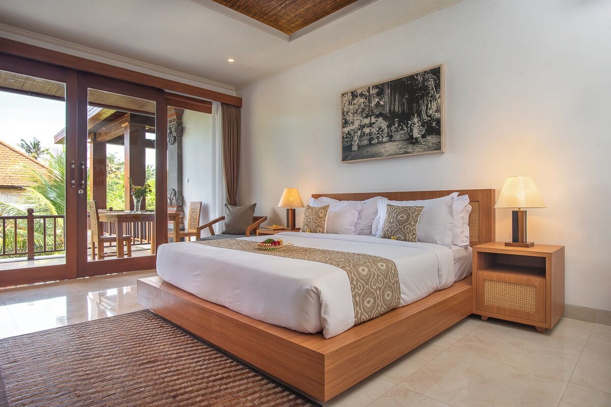 A spacious bedroom with a large bed, two bedside tables with lamps, a wooden headboard, and a large window with a view of greenery.