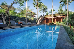 A swimming pool with lounge chairs and umbrellas, surrounded by palm trees and a wooden building. - Budhi Ayu Suite (Ubud)