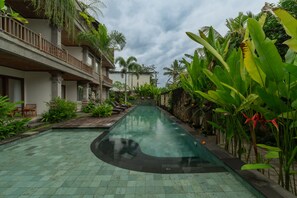 A swimming pool with a tiled deck, surrounded by lush greenery and a multi-story building with balconies. - Budhi Ayu Suite (Ubud)