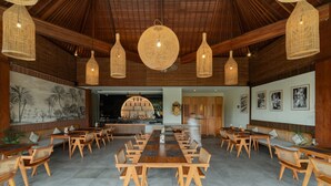 A spacious dining area with wooden tables and chairs, a large mural on the wall, and hanging wicker lamps. - Budhi Ayu Suite (Ubud)