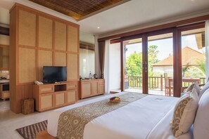 A bedroom with a large bed, a TV, a wooden wardrobe, and a balcony with a view of greenery. - Budhi Ayu Suite (Ubud)