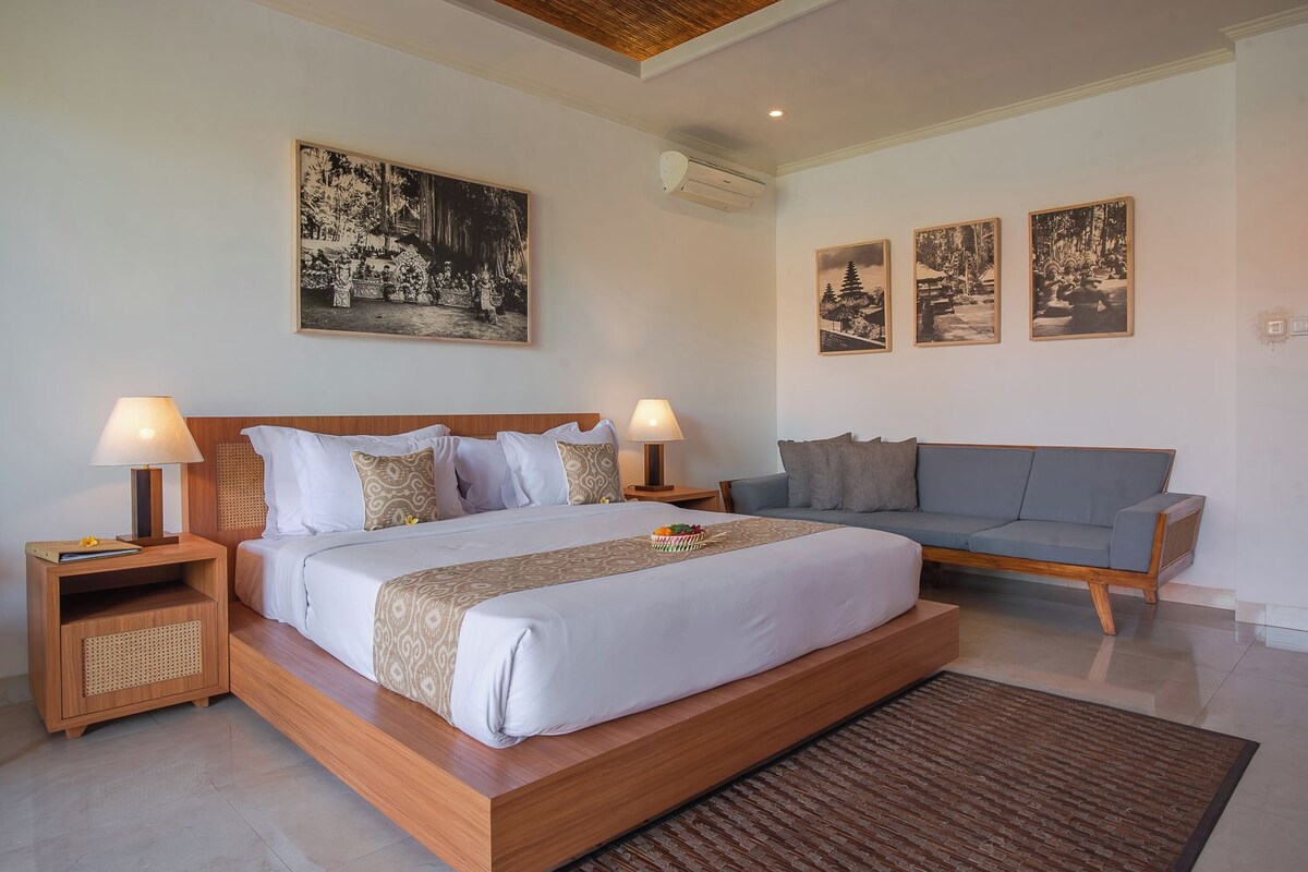 Une chambre moderne avec un grand lit, deux tables de chevet avec des lampes, un canapé gris et trois tableaux encadrés accrochés au mur.