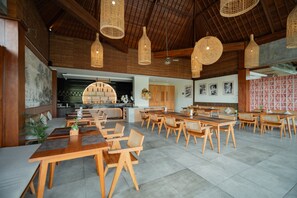 A spacious dining area with wooden tables and chairs, hanging wicker light fixtures, and a view into the kitchen area. - Budhi Ayu Suite (Ubud)