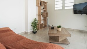 A modern living room with a wooden bookshelf, a TV mounted on the wall, and a large window.
