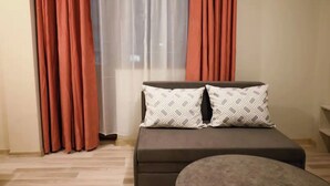 A bedroom with a bed, two patterned pillows, a small round ottoman, and a window with sheer curtains. - Hotel Leda (Sofia)