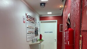 Un pasillo con pared roja, una puerta blanca que dice "ENTRADA SECRETA PARA FUMIAR", un extintor y un bote de basura.