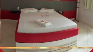 Una cama con cabecera roja y blanca, una mesita de noche con un teléfono, y una pared con interruptor de luz y toma de corriente.