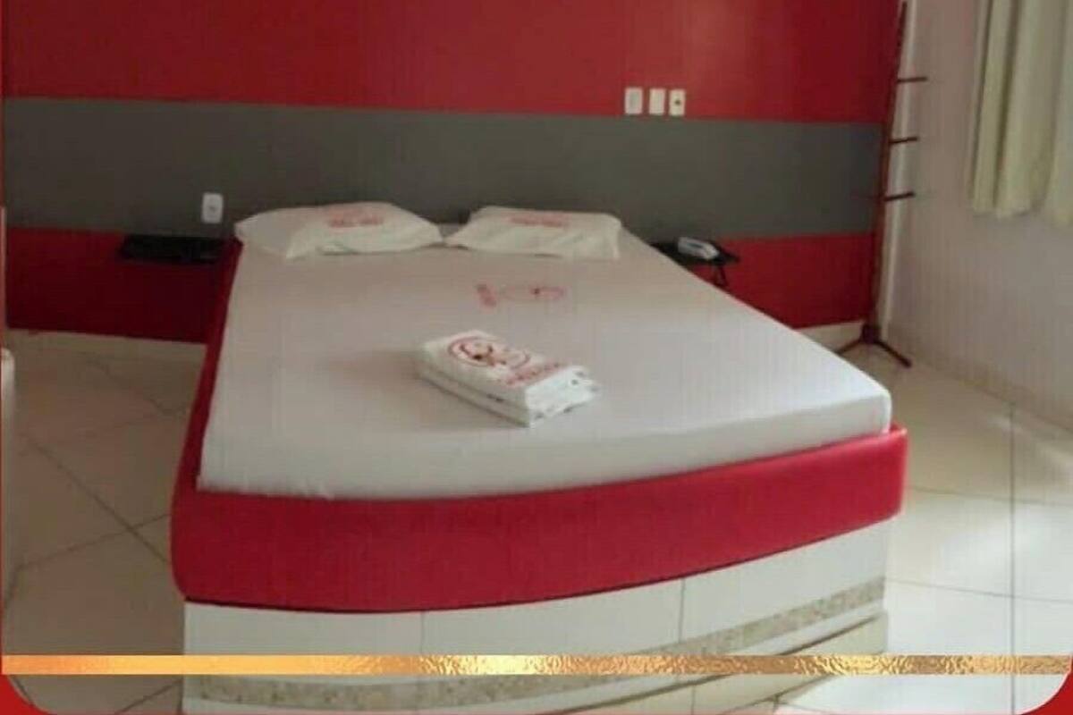 Un letto con una testiera rossa e bianca, un comodino con un telefono, e una parete con interruttore della luce e presa elettrica.