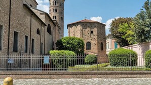 Un edificio histórico con una torre y una estructura adyacente más pequeña, rodeado de un área pavimentada y vegetación.