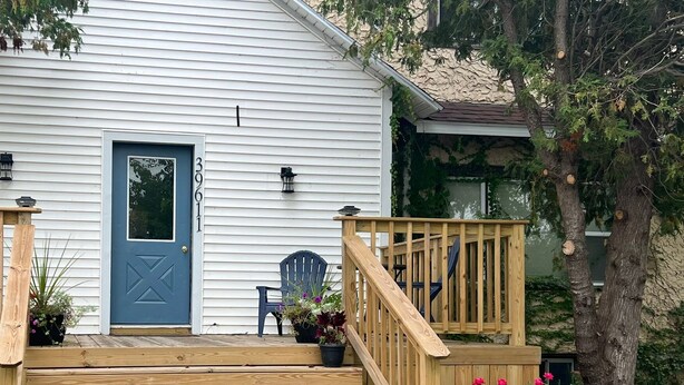 Una casa blanca con puerta azul, un porche delantero de madera y dos sillas negras.