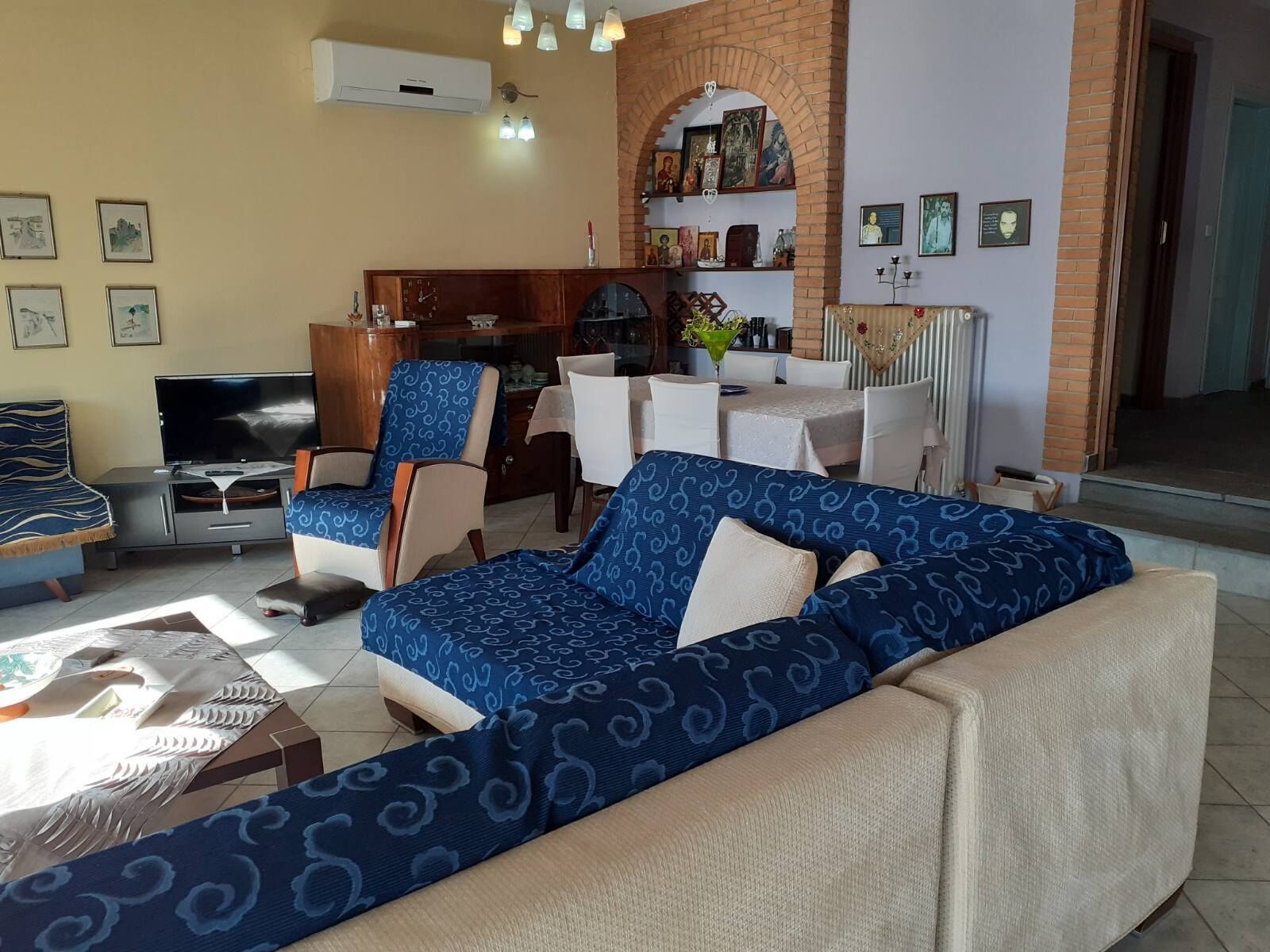 Uma sala de estar com um sofá azul estampado, uma televisão, um espaço de jantar com mesa e cadeiras, e uma parede de tijolo decorada com arte religiosa.