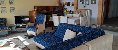 Uma sala de estar com um sofá azul estampado, uma televisão, um espaço de jantar com mesa e cadeiras, e uma parede de tijolo decorada com arte religiosa.