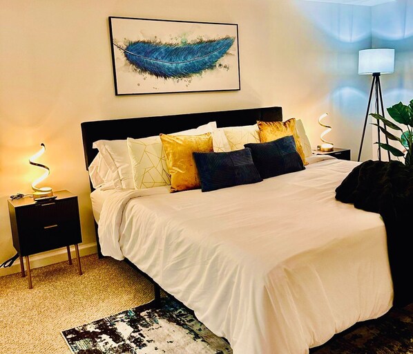 A neatly made bed with white bedding and multiple pillows, a bedside lamp, a framed artwork on the wall, and a floor lamp in the corner.