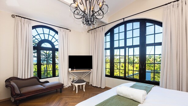 Ein Hotelzimmer mit einem großen Bett, einer Couch, einem Fernseher und zwei großen Fenstern mit Vorhängen.