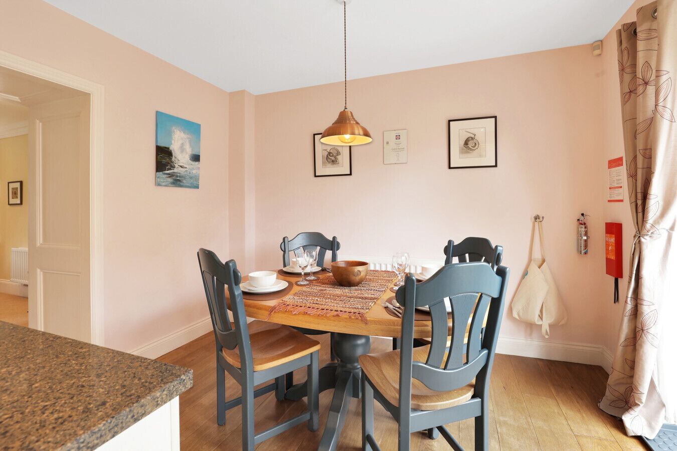 Uma área de refeições com mesa de madeira, quatro cadeiras, luminária pendente e uma moldura na parede.