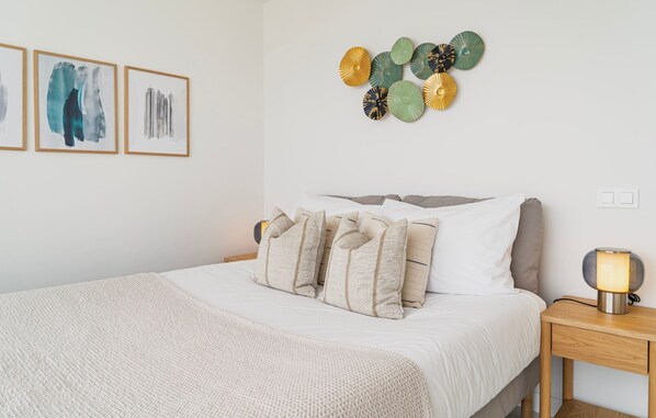 Un dormitorio ordenado con cama, almohadas, una mesita de noche con lámpara y decoración en la pared.
