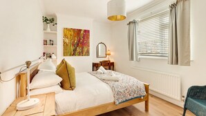 A bedroom with a wooden bed, a bedside table with a lamp, a bedside plant, a window with curtains, and a radiator. - Cherry Tree House- North East Escapes (Amble)
