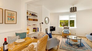 A cozy living room with a wooden dining table, wicker chairs, a sofa, a TV, and a bookshelf filled with books and decorative items. - Cherry Tree House- North East Escapes (Amble)