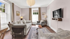 Een woonkamer met een bank, fauteuil en salontafel, een aan de muur bevestigde televisie, en een groot raam met gordijnen.