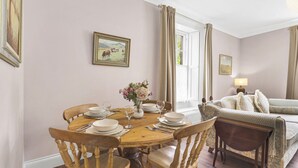 Een eetruimte met een ronde houten tafel voor vier personen, omringd door houten stoelen. Op de tafel staat een vaas met bloemen, en erboven hangt een schilderij aan de muur.