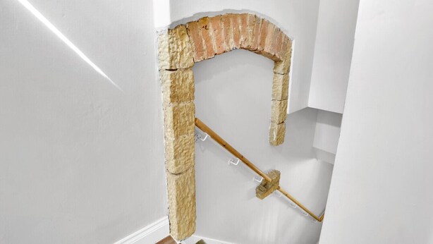 Een hoek van een kamer met een witte muur, een bakstenen boog en een houten bezem die tegen de muur leunt.