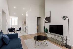 Una sala de estar moderna con un sofá azul, una mesa de centro de vidrio, un televisor sobre un mueble bajo y una cocina al fondo.