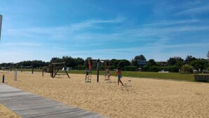 Zona arenosa de playa con un paseo marítimo de madera, una red de voleibol y algunas personas.