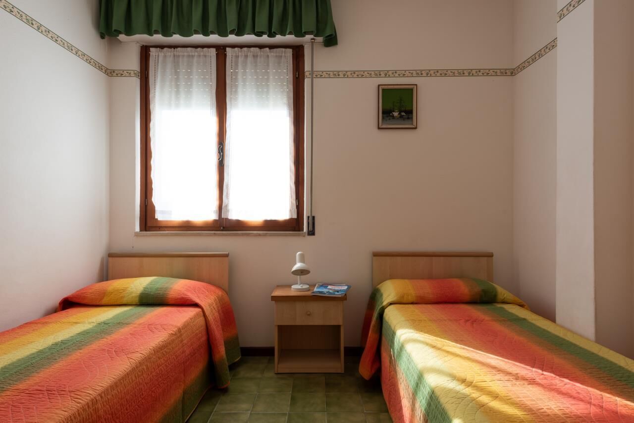 Una habitación pequeña y bien iluminada con dos camas individuales, una mesita de noche con lámpara y una ventana con cortina.